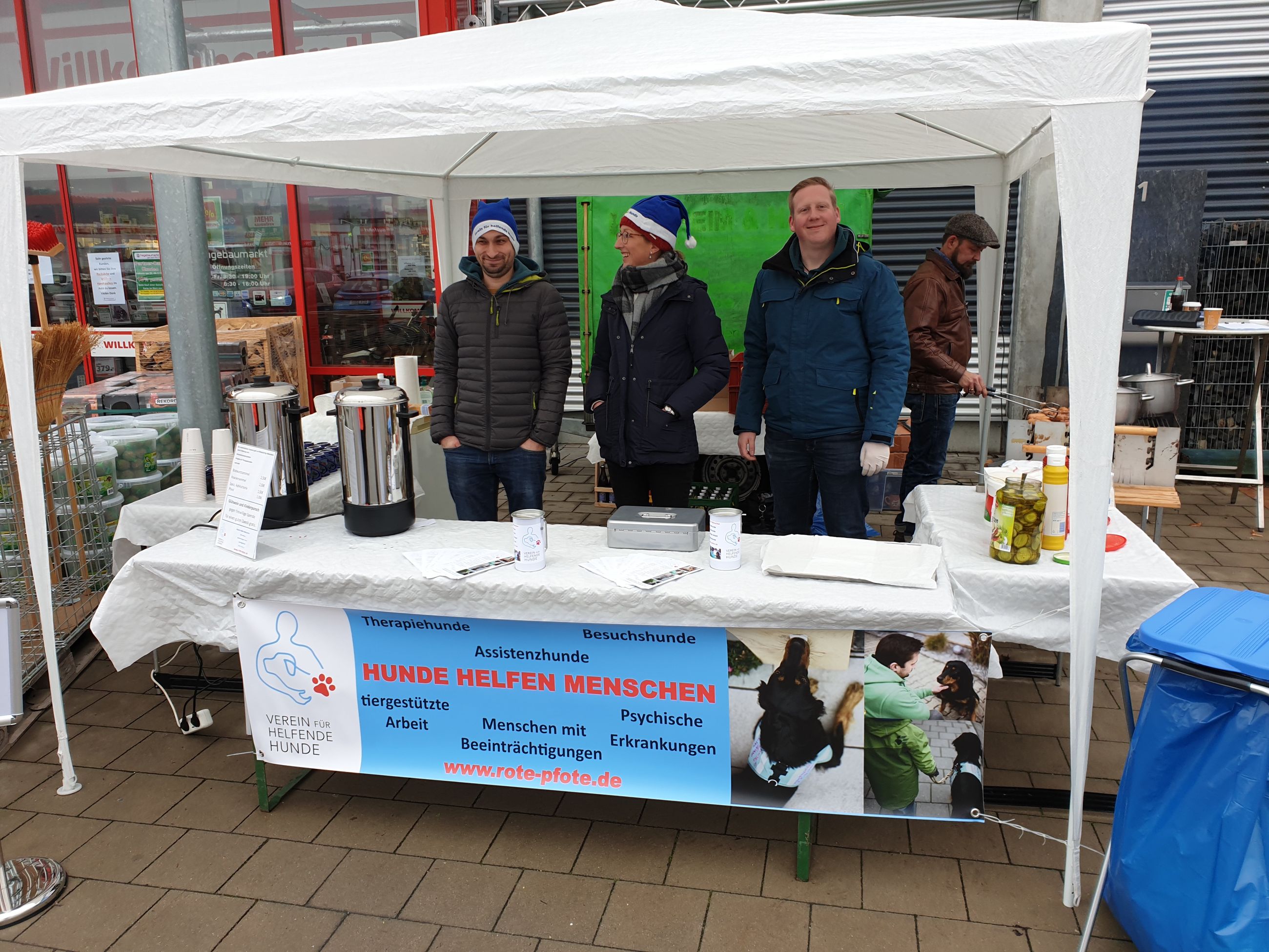 Informationspavillon des Vereins für helfende Hunde vor einem Baumarkt. 4 Menschen stehen unter einem Pavillon, hinter Stehtischen. 2 tragen blaue Weihnachtsmannmützen, eine Person im Hintergrund grillt.